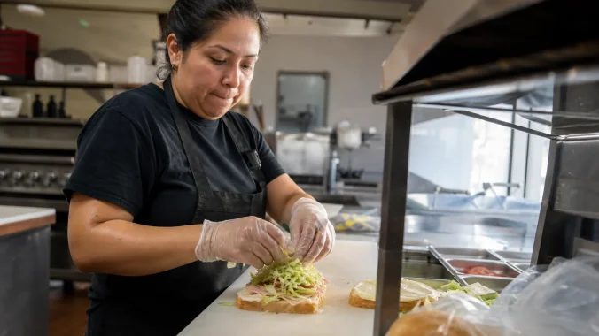 Worker wearing clear disposable gloves assembling sandwiches at a commercial kitchen deli station