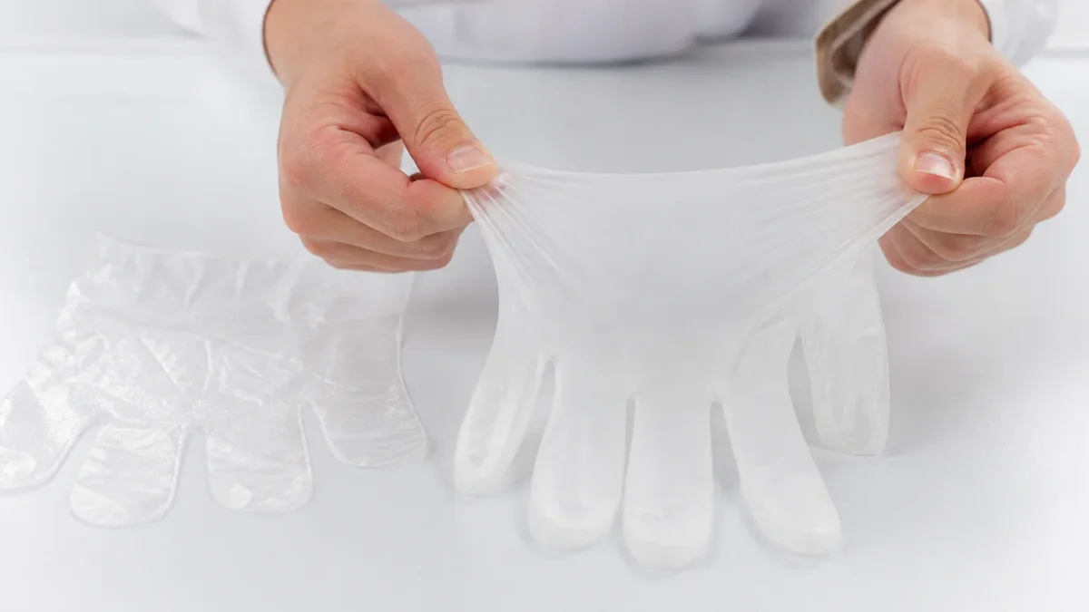 Close-up of a TPE glove being stretched to show elasticity beside a flat rigid PE glove on a white surface